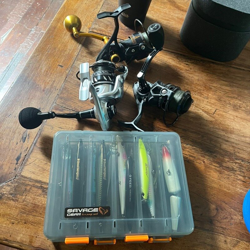 A Savage Gear lure box and three sea fishing reels on a wooden table