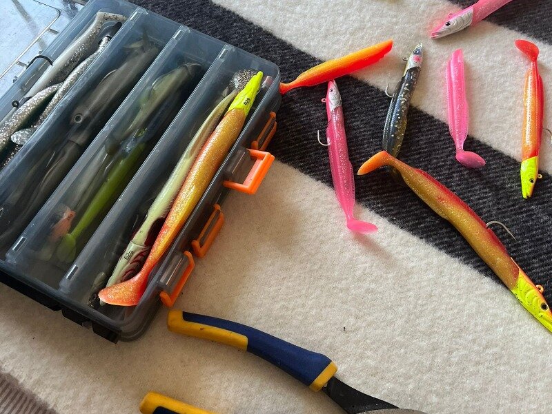 A classic pollock fishing lure box full of coloured lures on the bed next to some pliers